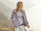 Een vrouw in een paarse jas en een T-shirt met print staat in een bergachtig landschap.