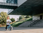 Een jongetje rijdt op een blauwe loopfiets voor een modern gebouw.
