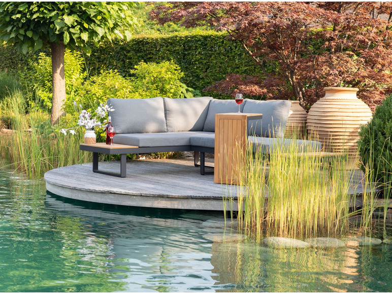 Een grijze hoekbank met een houten salontafel op een houten terras aan een vijver.