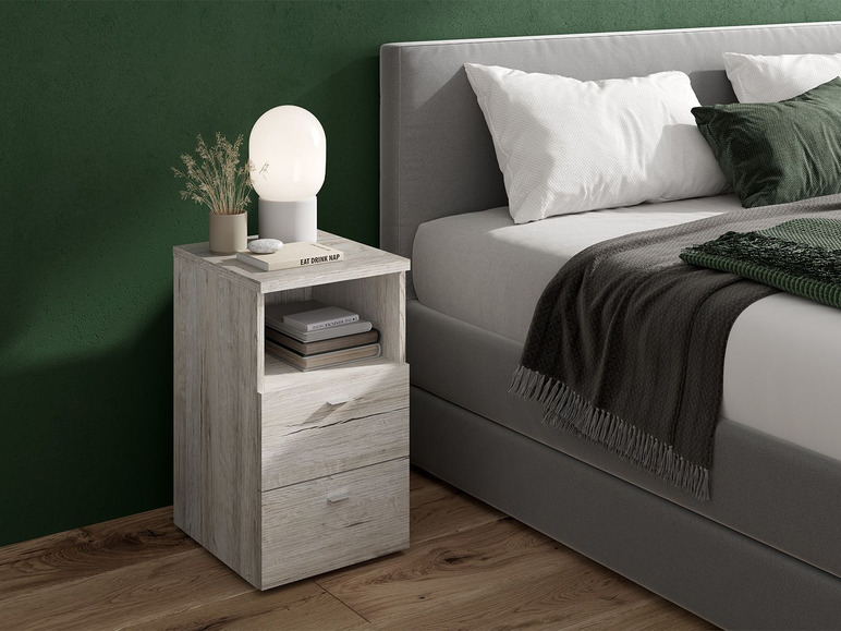 Een wit houten nachtkastje met twee laden naast een grijs bed.