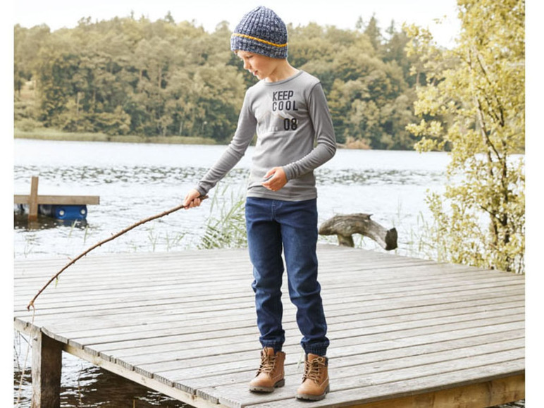 Jongetje met een grijze trui, blauwe jeans en een blauwe muts staat op een houten vlonder aan het water.