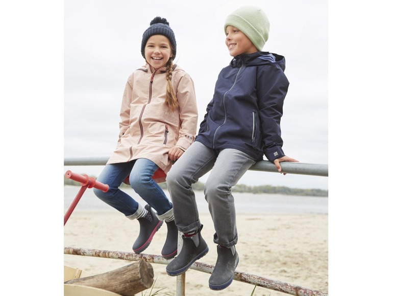 Twee kinderen in regenjassen en laarzen zitten op een hek.