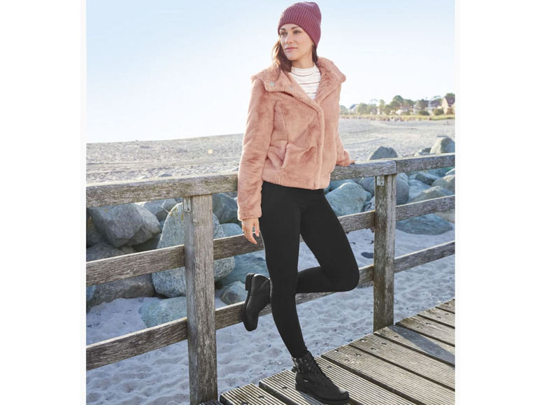 Een vrouw draagt een roze faux fur jas, zwarte leggings en zwarte laarzen.