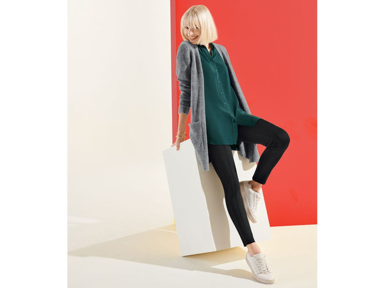 Vrouw in een grijs vest, een groene blouse en zwarte leggings.