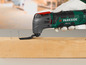 Een groene Parkside multitool met een mesje, gebruikt om een houten plank te bewerken.