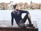 Man in een beanie, trui en jeans zit op een houten pier bij een meer.