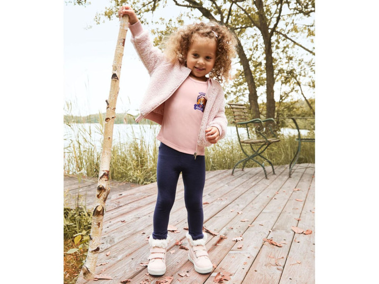 Een meisje draagt een roze trui met Paw Patrol print, blauwe leggings en witte sneakers.