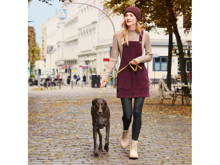 Een vrouw in een donkerrode jurk en zwarte panty's loopt met haar hond over straat.