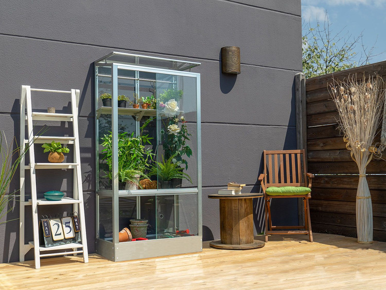 Een glazen kas met planten en een houten stoel met een groene kussen op een terras.