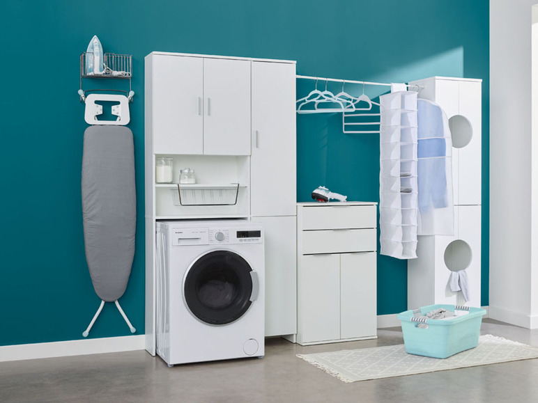 Een wasmachine, een strijkplank en een wasmand in een witte wasruimte.