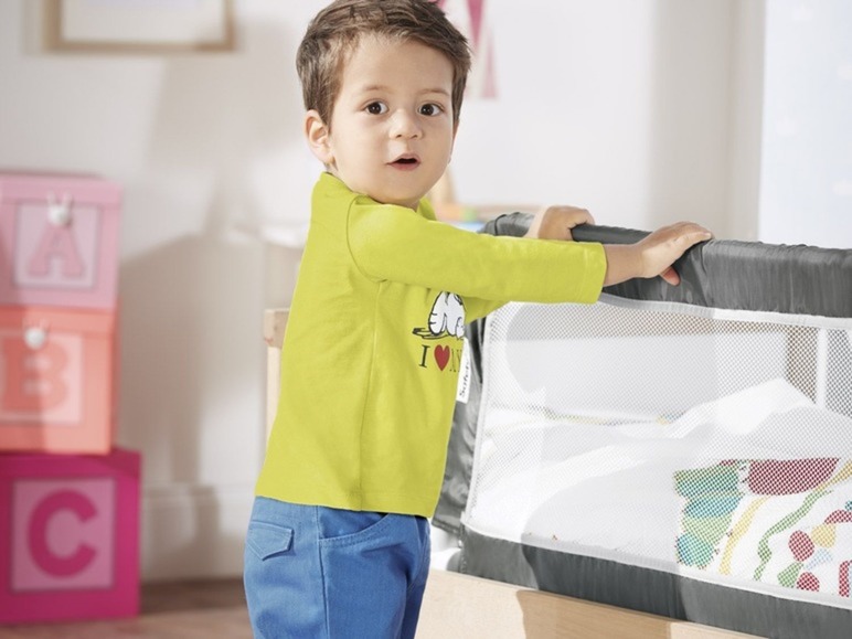 Een kind in een geel shirt en blauwe broek houdt zich vast aan een bed met een veiligheidsscherm.