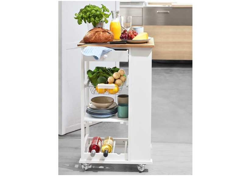 Een witte keukentrolley met een houten werkblad, gevuld met fruit, groenten, wijn en servies.