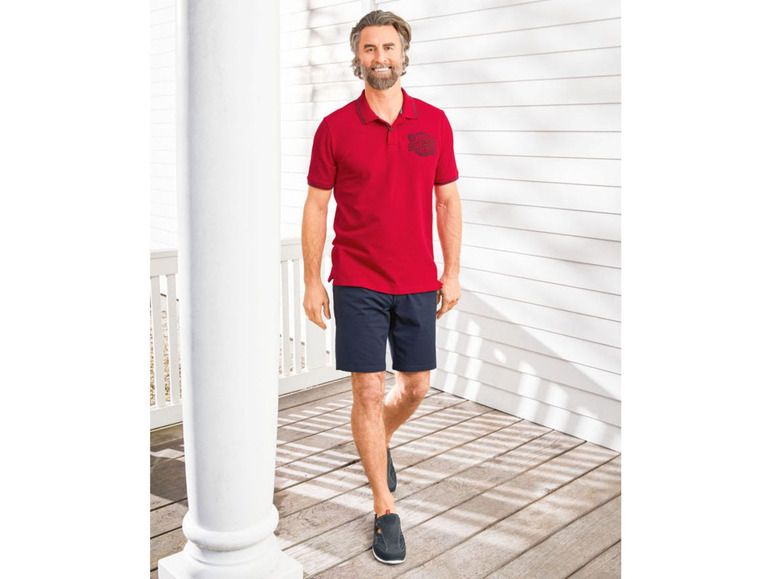 Man in een rode polo met korte mouwen, blauwe korte broek en zwarte schoenen.