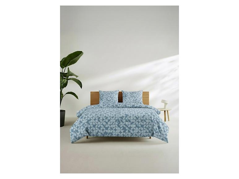 Slaapkamer met blauw beddengoed met patroon, houten hoofdbord, plant en bijzettafel.