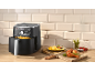 Zwarte airfryer met friet, kippenpoten en tomaten op een houten tafel.