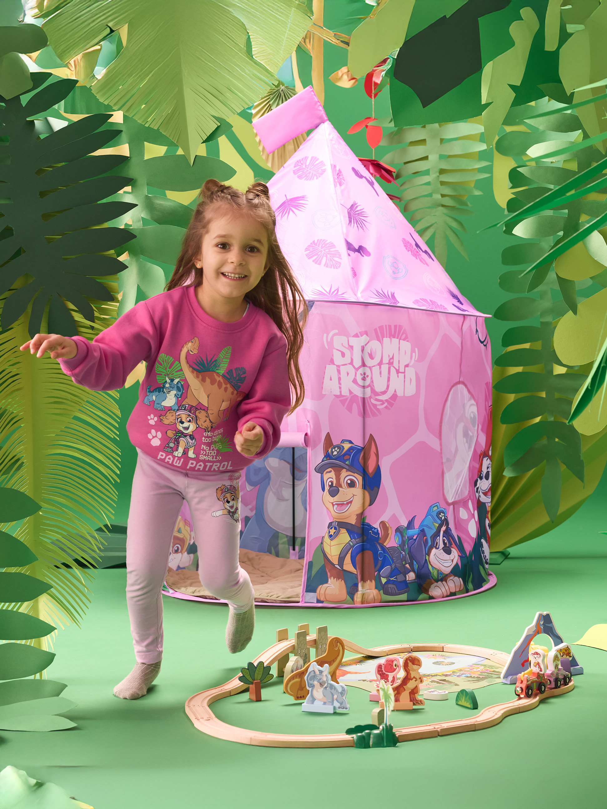 Meisje in roze sweatshirt en legging, spelend met een roze speeltent en een houten treinset.