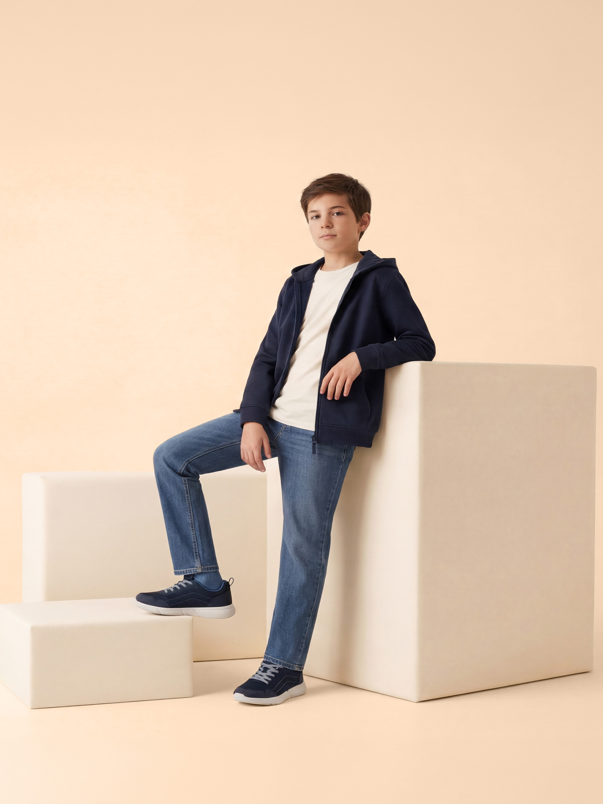 Jongen in blauwe hoodie, wit T-shirt en blauwe jeans, leunend op witte blokken.