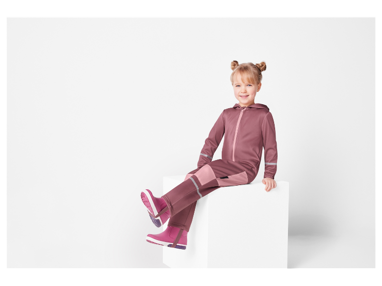 Een lachend meisje zit op een witte kubus, gekleed in een roze jumpsuit en roze laarzen.
