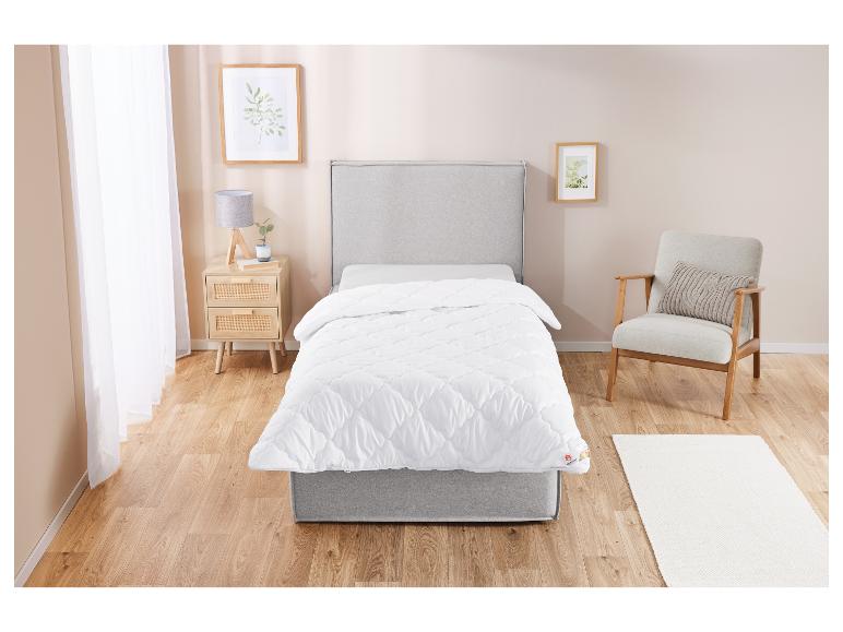 Een wit, gestikt dekbed op een grijs bed in een lichte slaapkamer.