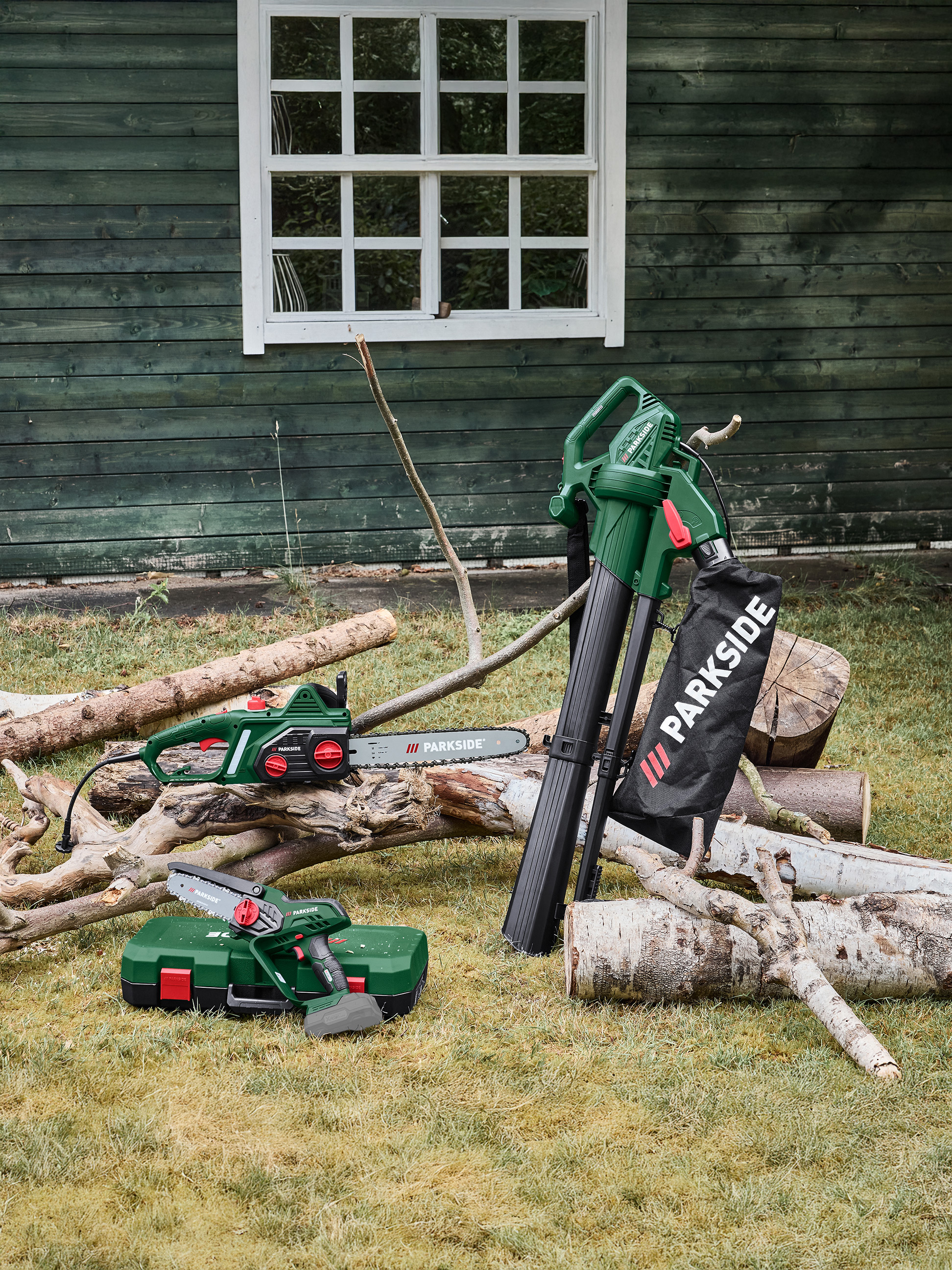 Bladblazer, kettingzaag en minikettingzaag op gras bij een houten schuur.
