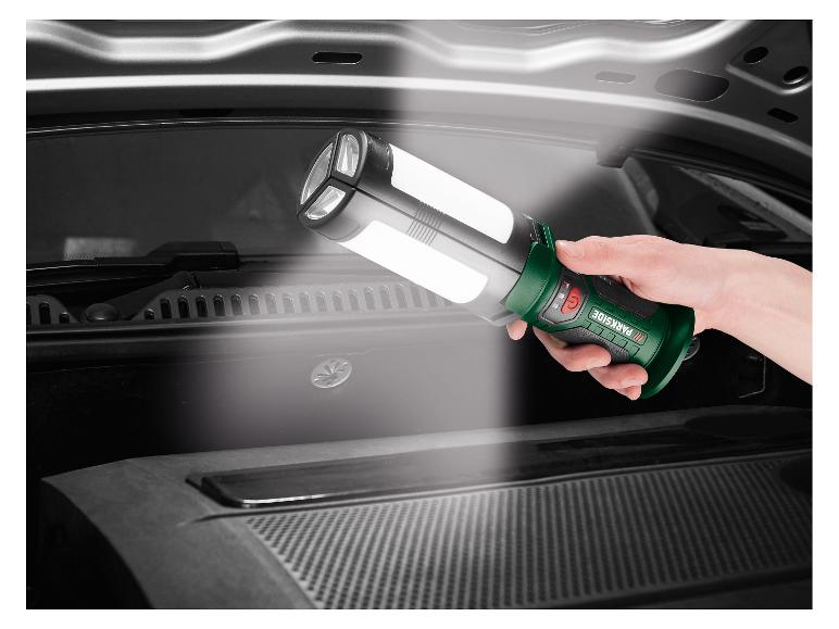 Een hand houdt een groene en zwarte werklamp vast, die de motorruimte van een auto verlicht.