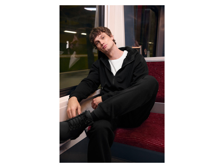 Man in een zwarte hoodie en broek, zit op een treinbank