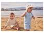 Twee baby's in biologisch katoenen strandkleding op het zand, met zee en stad op de achtergrond.