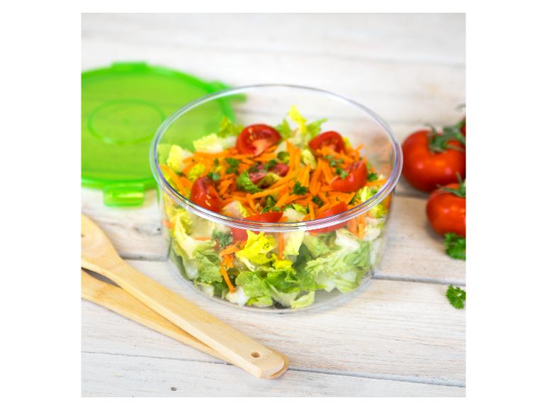 Een doorzichtige kom met salade, tomaten en wortels, met een groen deksel en houten keukengerei op een lichte houten tafel.
