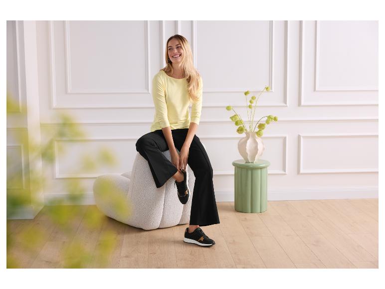 Vrouw in geel shirt en zwarte broek zit op een beige stoel.