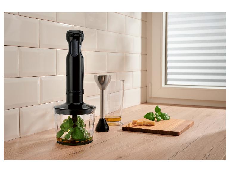 Staafmixer met maatbeker, basilicum, kaas en olijfolie op houten aanrecht.