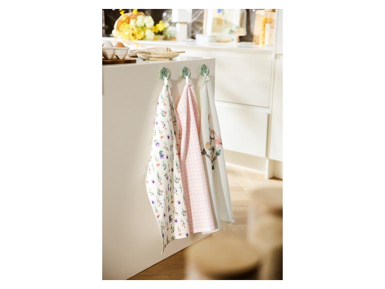 Drie theedoeken met bloemen- en ruitpatronen hangen aan haken in een keuken.