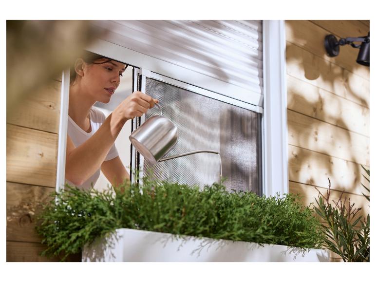 Vrouw giet planten in een bloembak voor een raam met rolluik en hor.