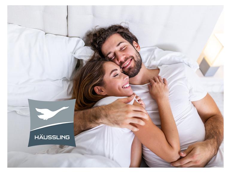 Lachend stel in bed met Häussling-logo op een kussen.