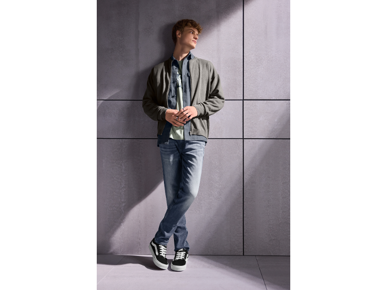 Jonge man in grijs jack, jeans en sneakers, leunend tegen een muur.