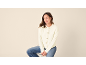 Vrouw in crèmekleurig vest en jeans zit op een kruk, kijkt naar rechts.