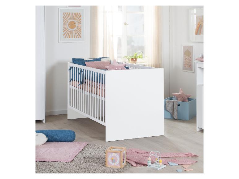 Wit babybedje met roze en blauwe kussens, speelgoed en zonnewanddecoratie.