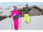 Twee kinderen in winterkleding met skistokken in een besneeuwd berglandschap met een houten hut.