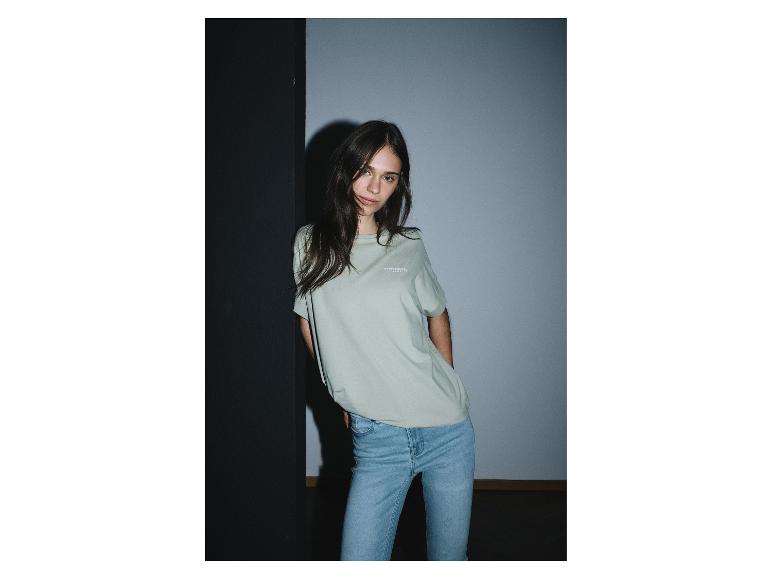 Een vrouw in een lichtgroen t-shirt met de tekst 'STRENESS BLUE' en lichtblauwe jeans.