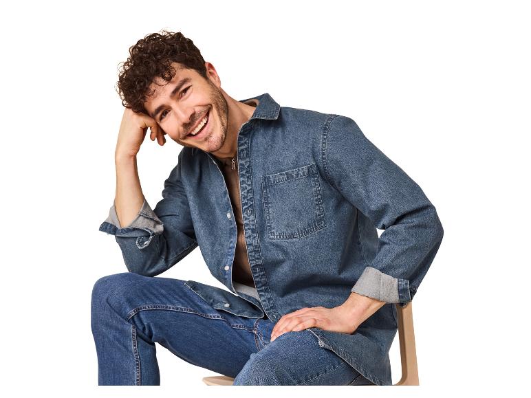 Lachende man in een spijkeroverhemd en jeans, zittend op een stoel.