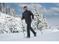 Man in zwarte winterjas en broek wandelt met stokken in besneeuwd boslandschap.
