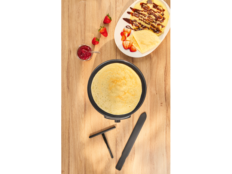 Pannenkoek in pan, ernaast bord met pannenkoeken, bananen, aardbeien en jam.