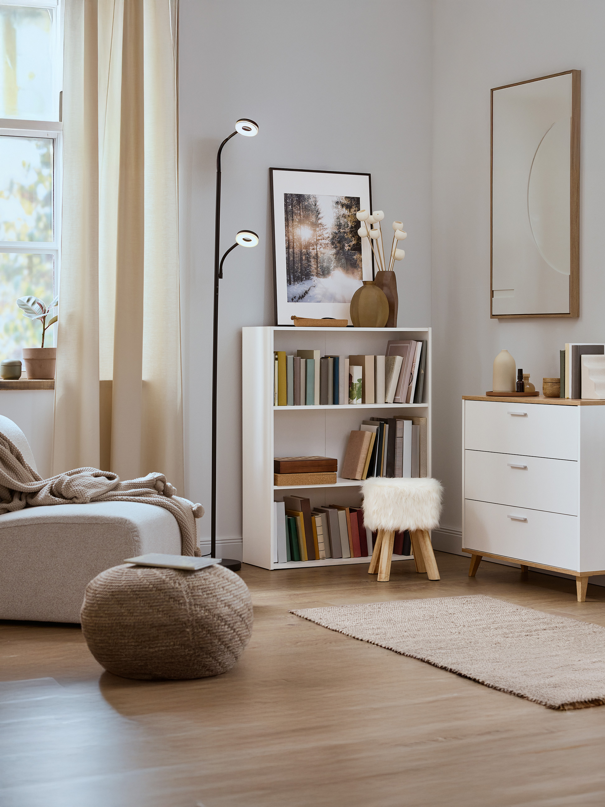 Lichte woonkamer met een witte boekenkast, ladekast, fauteuil en vloerlamp.