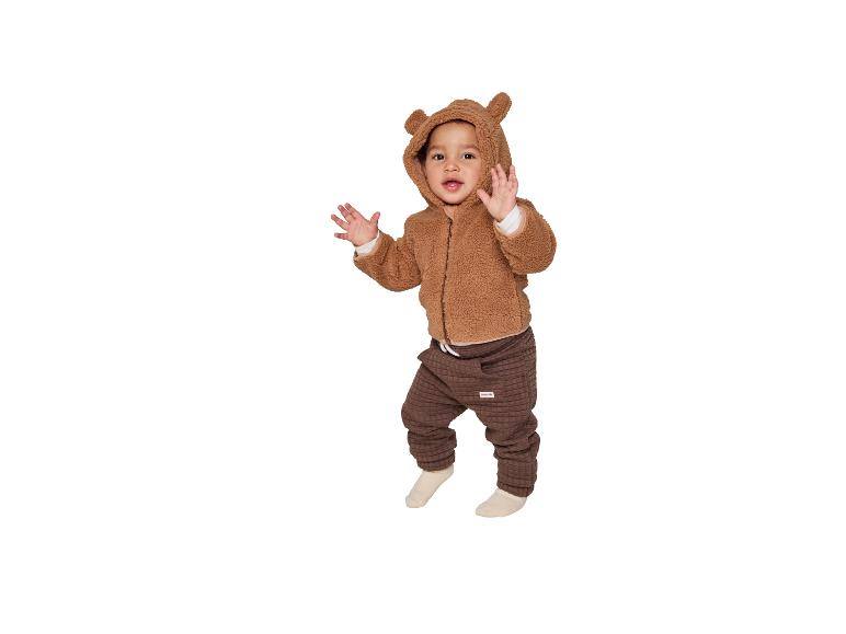 Baby in een bruine teddybeer hoodie en bruine broek, zwaaiend naar de camera.