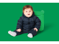 Baby in een donkerblauw gewatteerd skipak met berenembleem, zittend naast een groene speelgoeddoos.