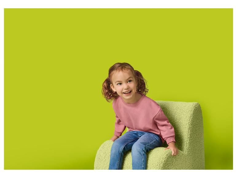 Lachend meisje in roze sweatshirt en jeans zit op een groene fauteuil
