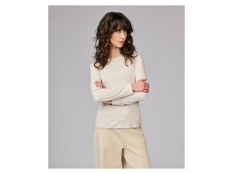 Vrouw in een beige geribbelde top met lange mouwen en beige broek, met gekruiste armen.
