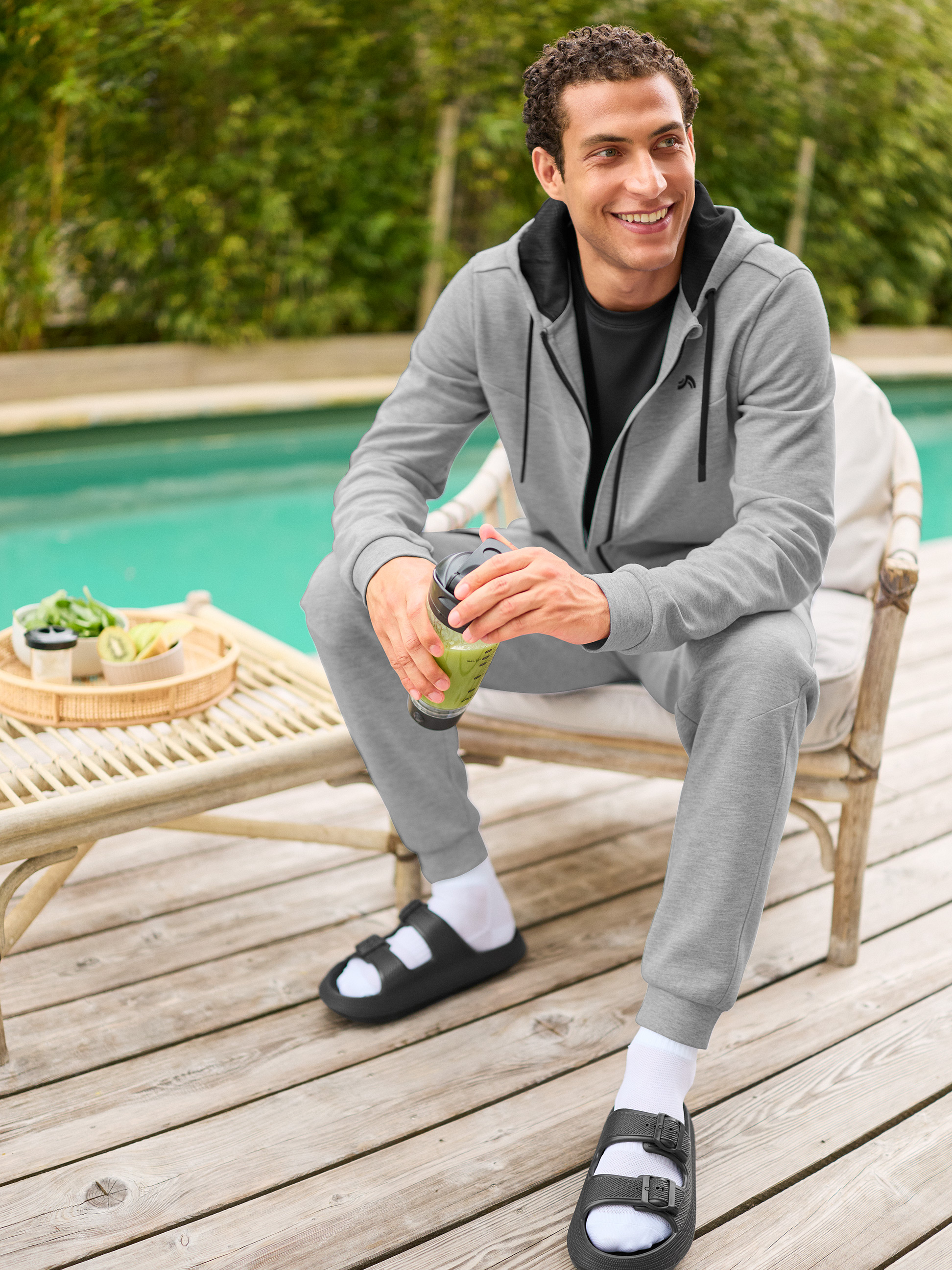 Man in grijs trainingspak en zwarte sandalen met witte sokken, met groene smoothie bij een zwembad.