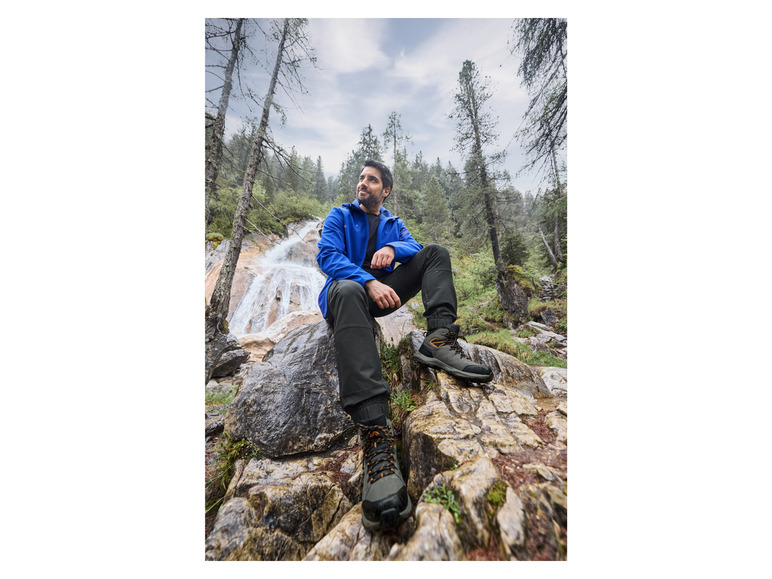 Een man in een blauwe jas en bruine wandelschoenen zit op rotsen in een bos.