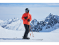 Man in oranje ski-jas, zwarte skibroek, helm en skibril op besneeuwde bergtop.