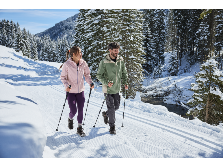 Twee mensen met wandelstokken lopen op een besneeuwd pad tussen sparren.